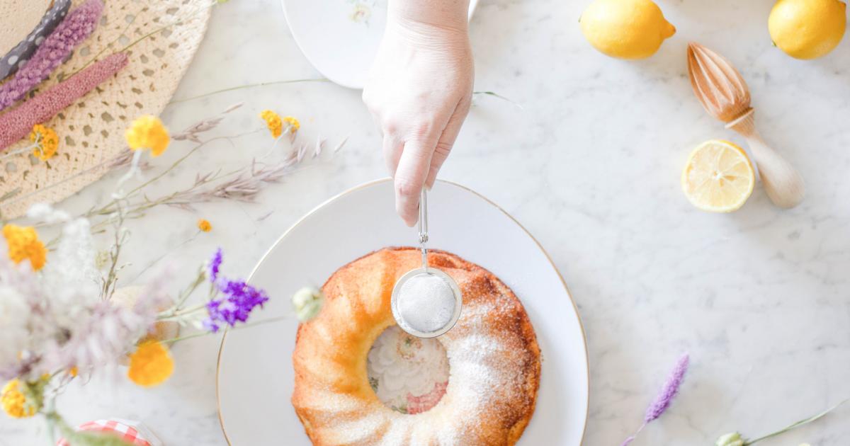 lemon bundt cake on white marble with white persons hand holding stainless steel sieve with icing sugar, lemons and flowers next to the cake too